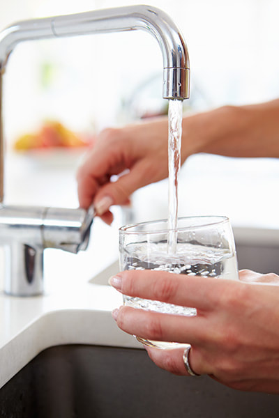 Photo of a woman filling a water glass with municipal drinking water from a kitchen sink faucet.